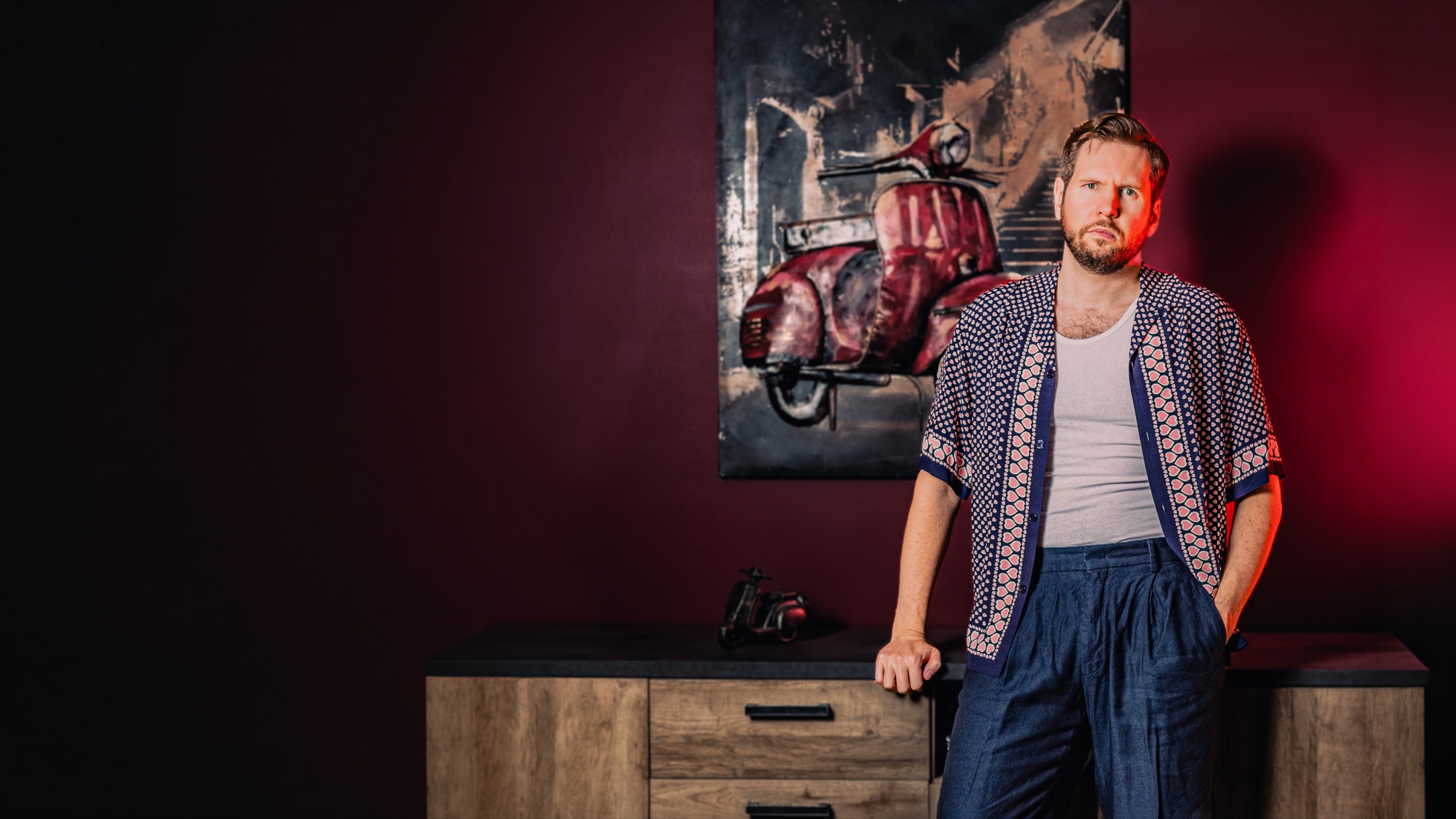 Sonny Zamolo devant un mur bordeaux et un tableau de scooter, portrait éclairé en rouge.